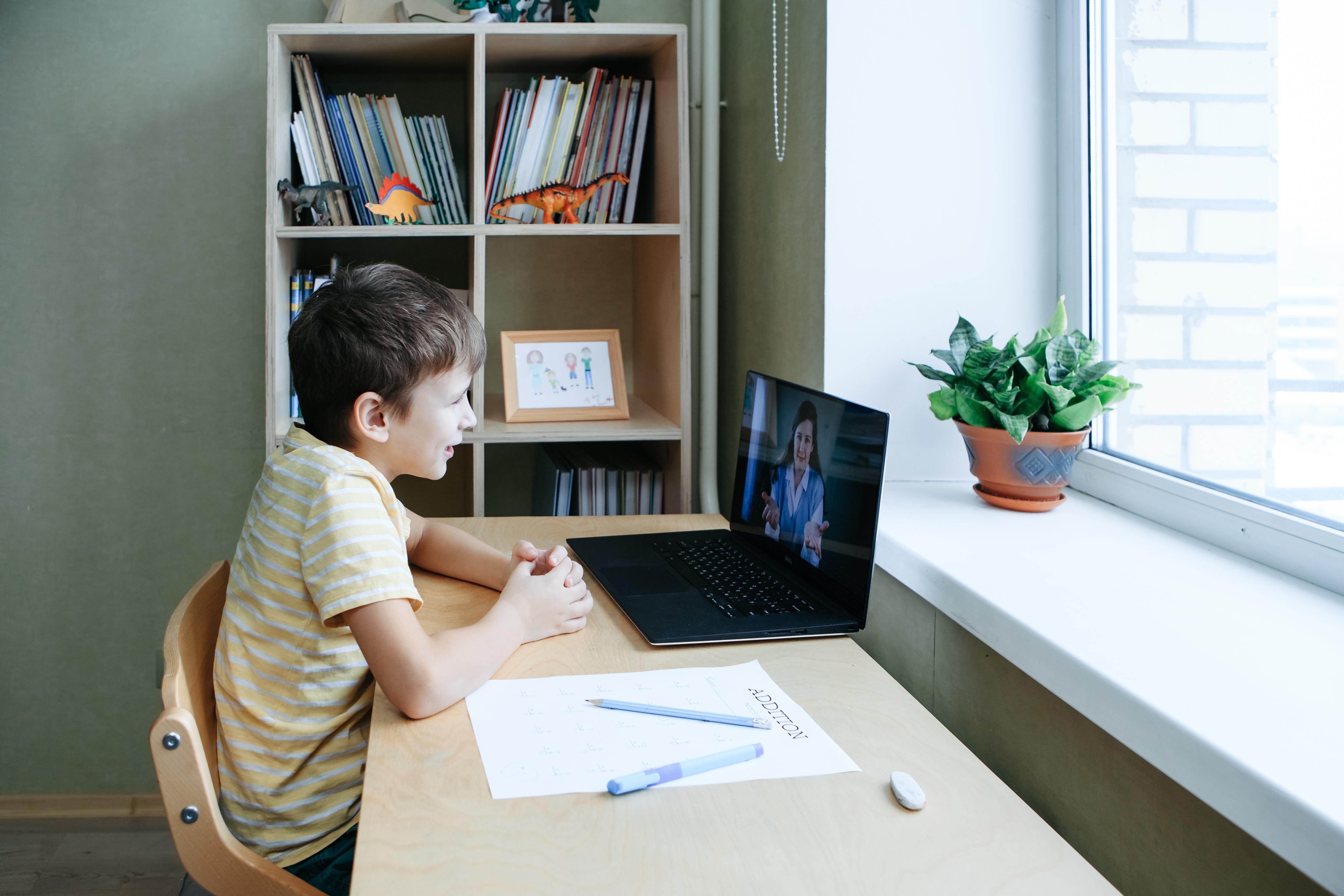 Young boy sitting at a table in front of a window, talking to a tutor on a laptop open in front of him