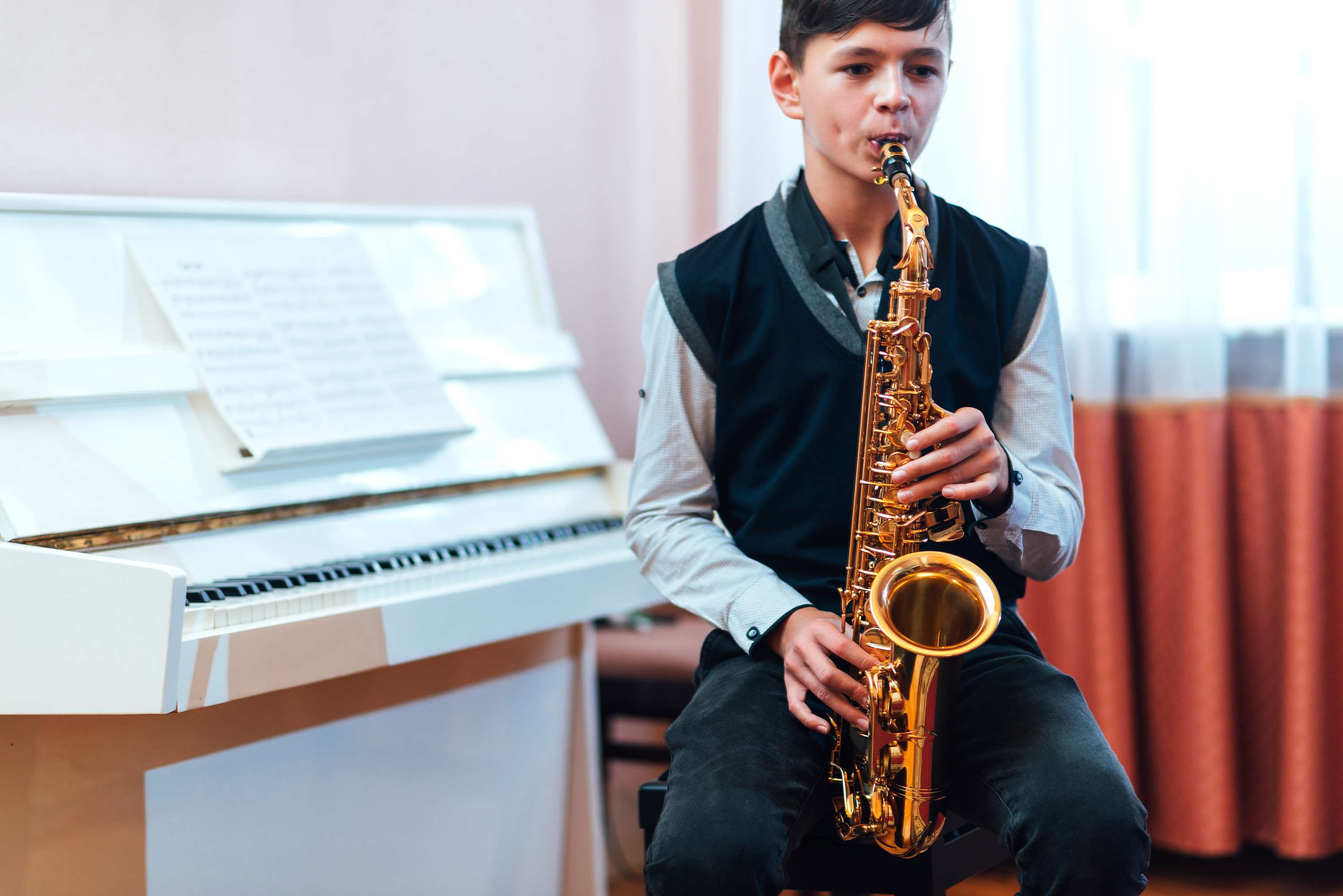 Boy in school uniform playing a saxophone while sitting at a piano in a music room