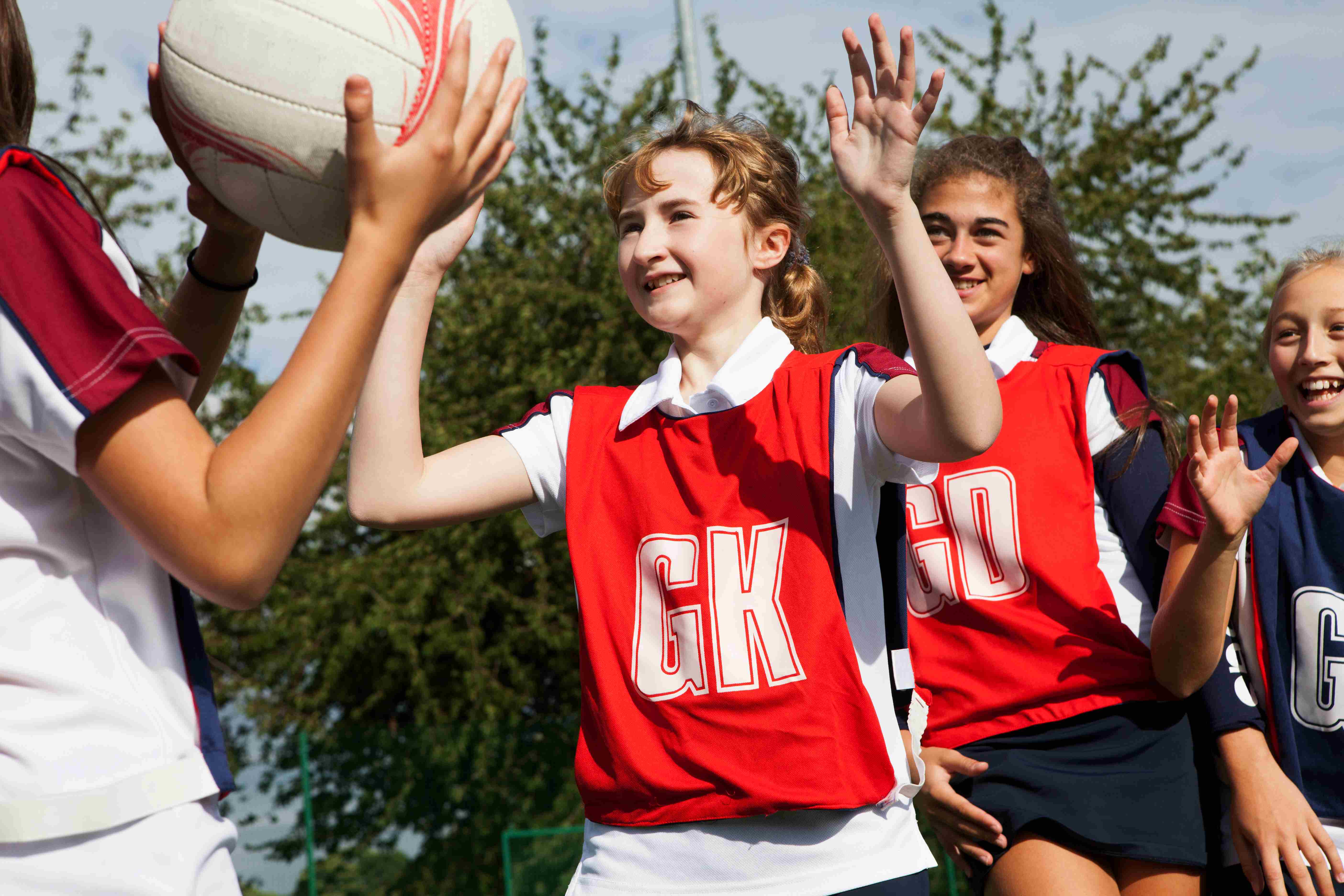 Smiling girls playing netball as part of a school team. The girl at the centre of the picture is wearing a goalkeeper's vest and is marking a player with the ball.